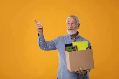 Happy unemployed senior man with box of personal office belongings on orange background. Space for text