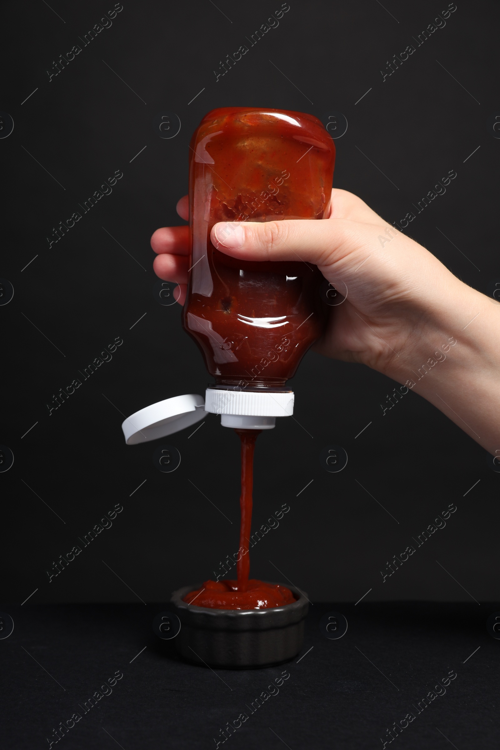 Photo of Woman pouring tasty ketchup from bottle into bowl against black background, closeup