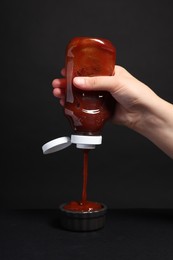 Photo of Woman pouring tasty ketchup from bottle into bowl against black background, closeup