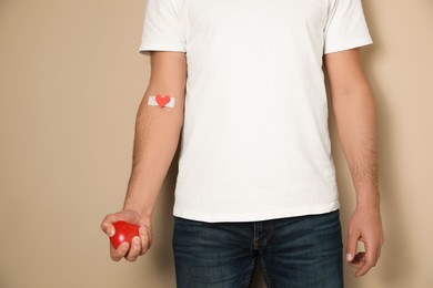Blood donation concept. Man with adhesive plaster on arm holding red heart against beige background, closeup