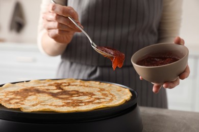 Photo of Woman cooking delicious crepe with jam on electric pancake maker in kitchen, closeup