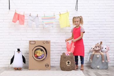 Little girl holding clean laundry near toy cardboard washing machine indoors