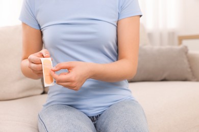 Photo of Woman opening sticking plaster on sofa indoors, closeup