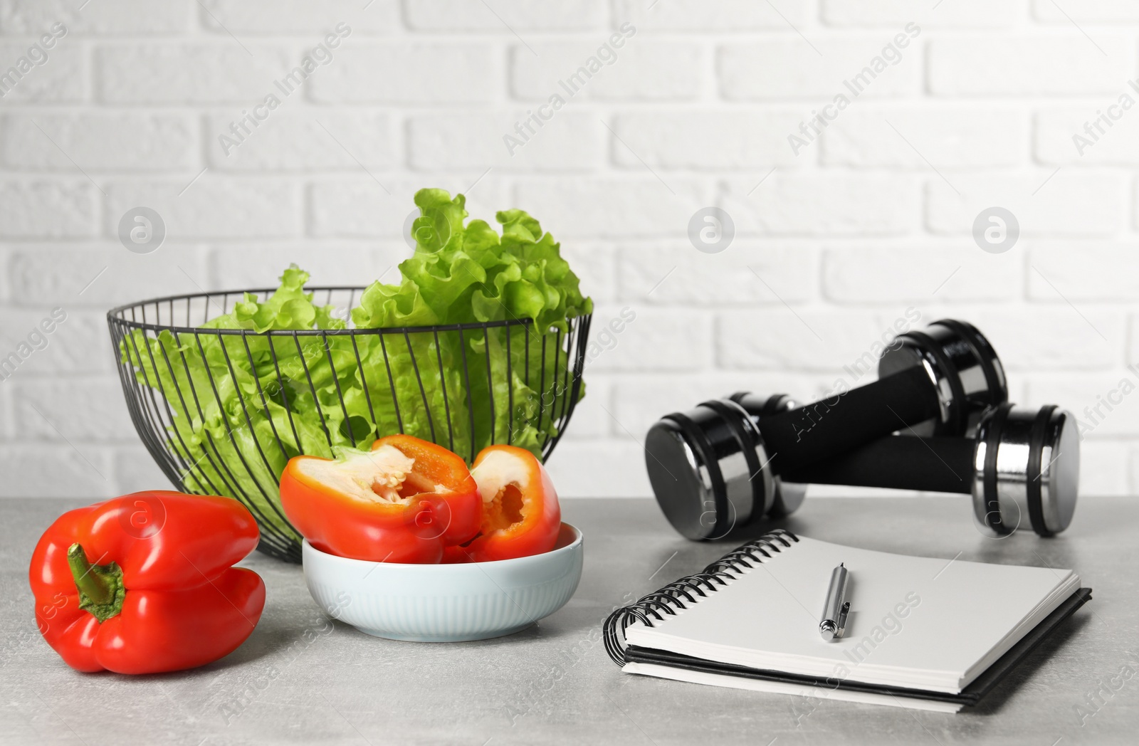 Photo of Healthy diet. Vegetables, dumbbells, notebook and pen on grey table