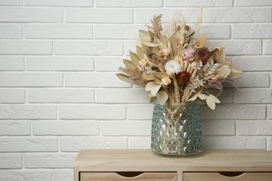 Beautiful dried flower bouquet in glass vase on wooden table near white brick wall. Space for text