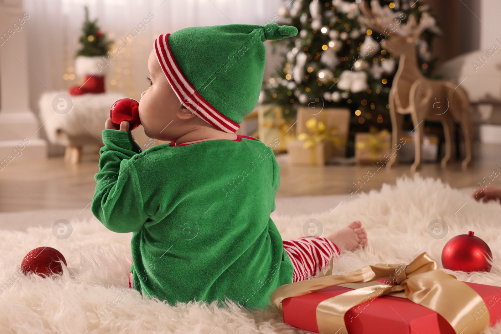 Photo of Baby in cute elf costume near Christmas gift on floor at home, back view