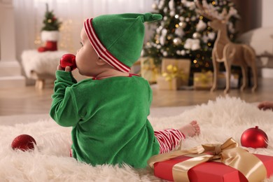 Baby in cute elf costume near Christmas gift on floor at home, back view