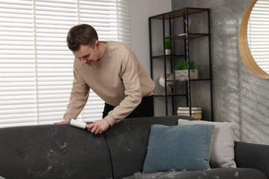 Photo of Pet shedding. Smiling man with lint roller removing dog's hair from sofa at home
