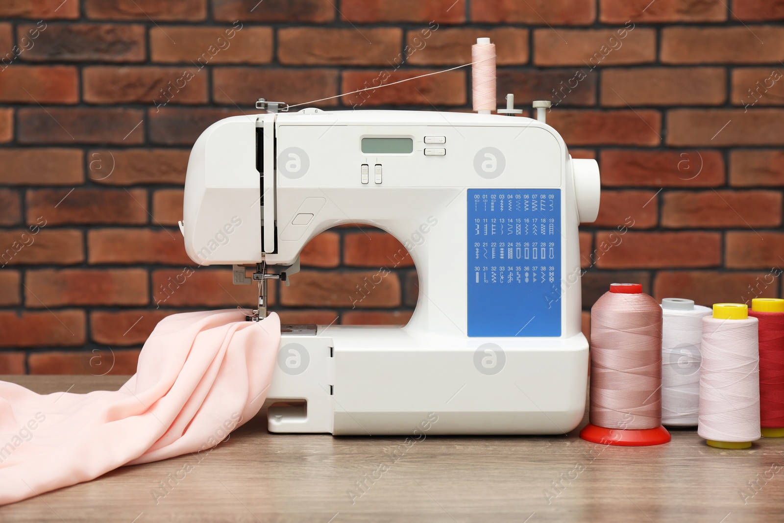 Photo of Sewing machine with fabric and spools of threads on wooden table against brick wall