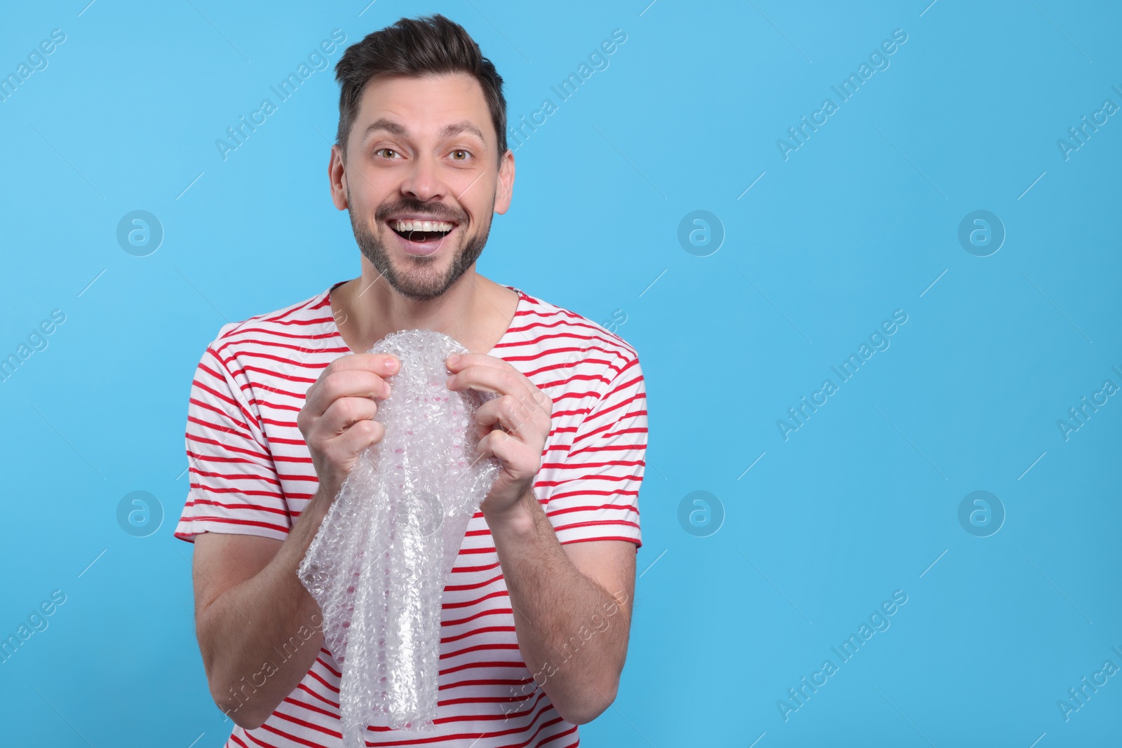 Photo of Happy man popping bubble wrap on light blue background, space for text. Stress relief