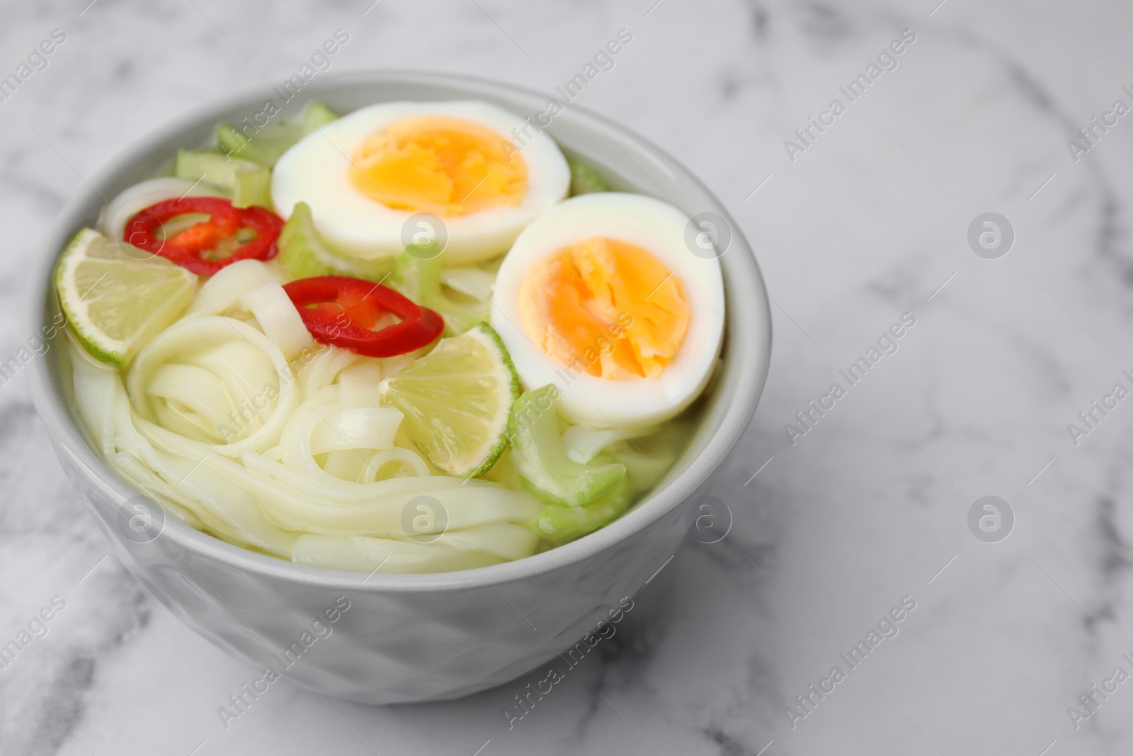 Photo of Bowl of delicious rice noodle soup with celery and egg on white marble table, closeup. Space for text