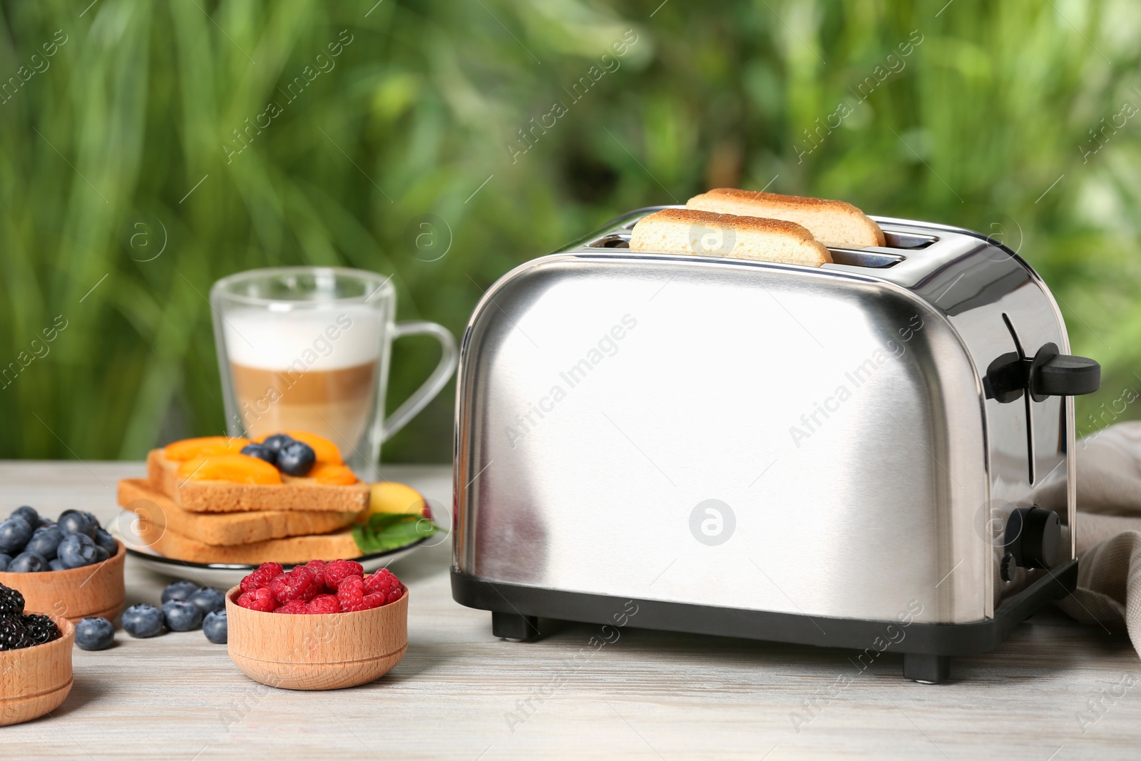 Photo of Modern toaster and fresh products on white wooden table against blurred green background