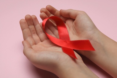 Photo of Little girl holding red ribbon on pink background, closeup. AIDS disease awareness