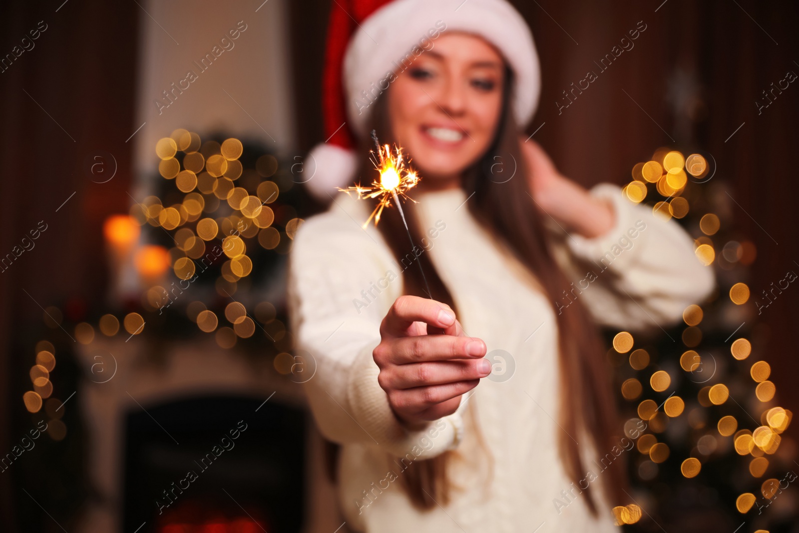 Photo of Beautiful young woman in Santa hat with burning sparkler indoors, focus on firework. Christmas celebration