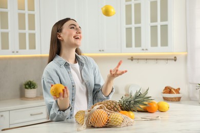 Photo of String bag with fresh fruits on light marble table. Woman throwing lemon in kitchen