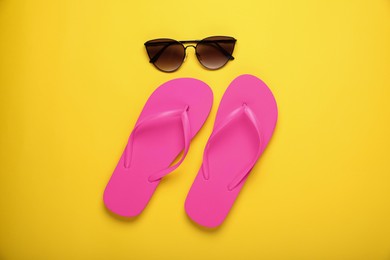 Photo of Stylish pink flip flops and sunglasses on yellow background, flat lay