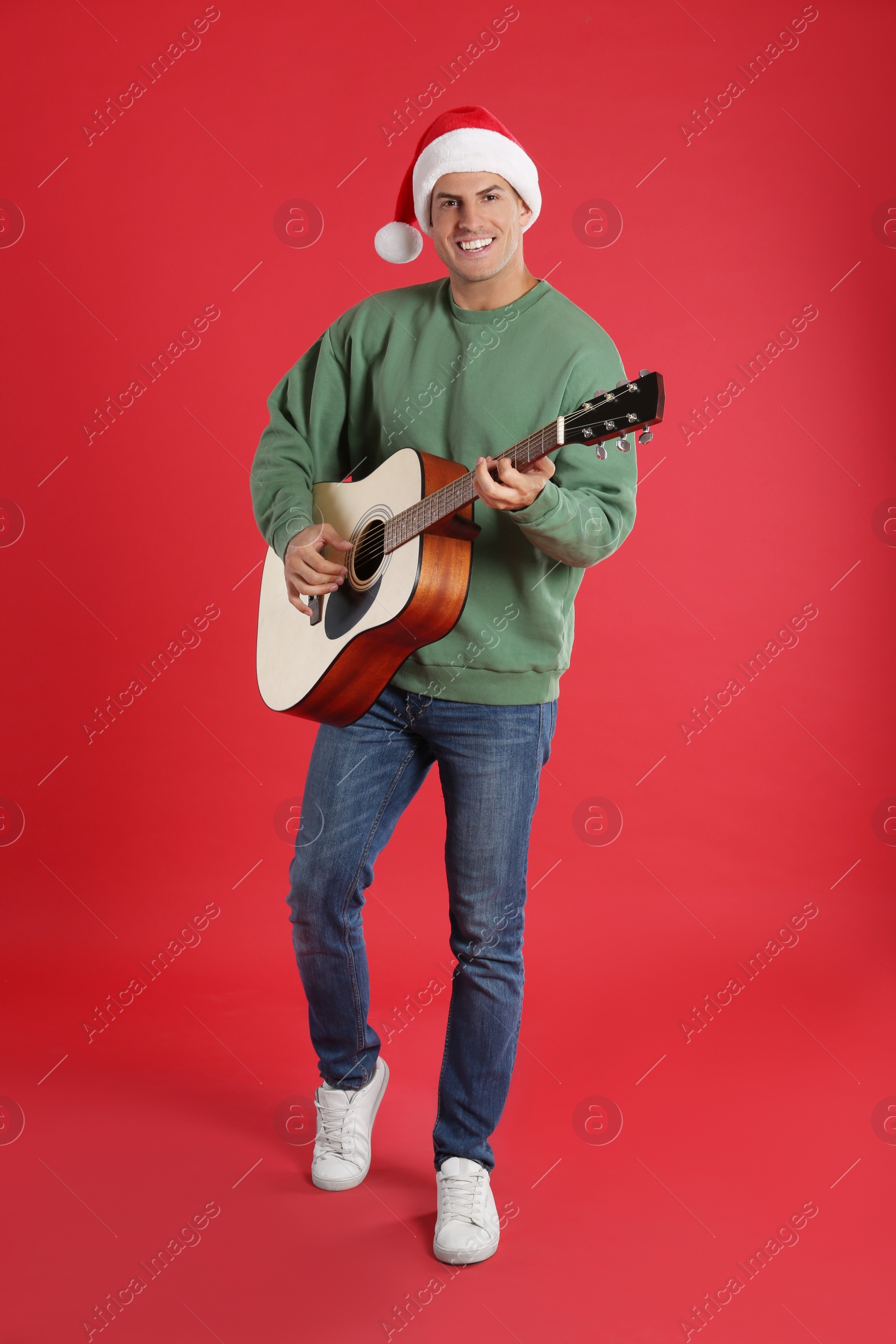Photo of Man in Santa hat playing acoustic guitar on red background. Christmas music