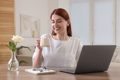 Photo of Happy woman with cup of drink near laptop at wooden table in room