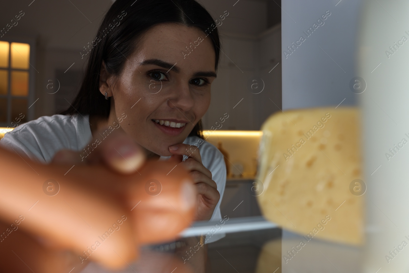 Photo of Young woman taking sausages out of refrigerator in kitchen at night, view from inside