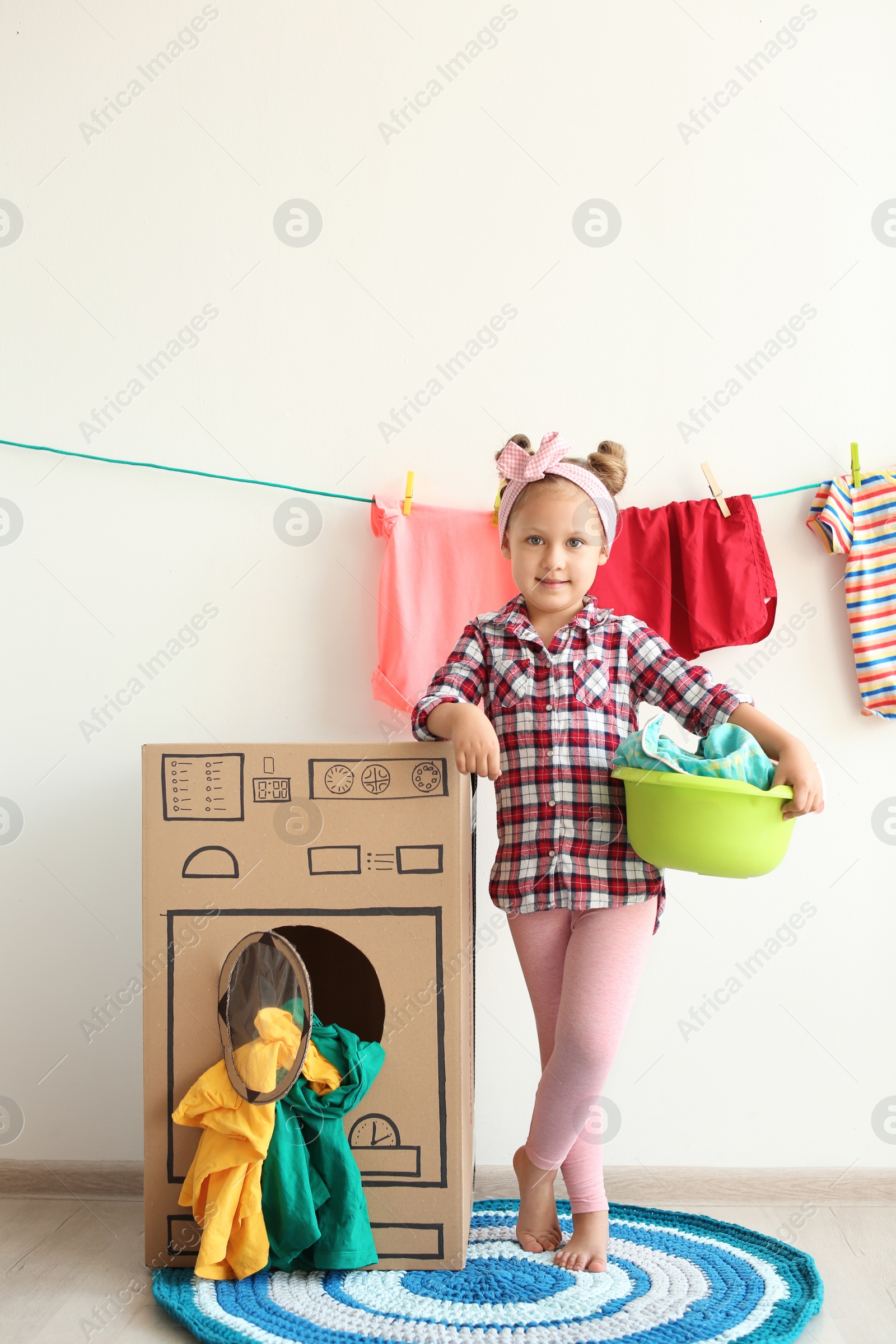 Photo of Adorable little child playing with cardboard washing machine and clothes indoors