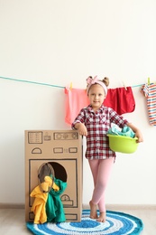 Photo of Adorable little child playing with cardboard washing machine and clothes indoors