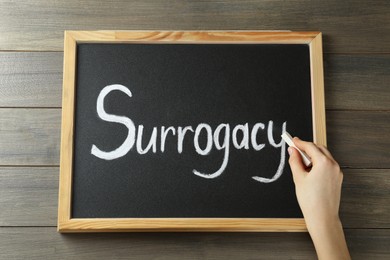 Woman writing word Surrogacy on small blackboard at wooden table, top view