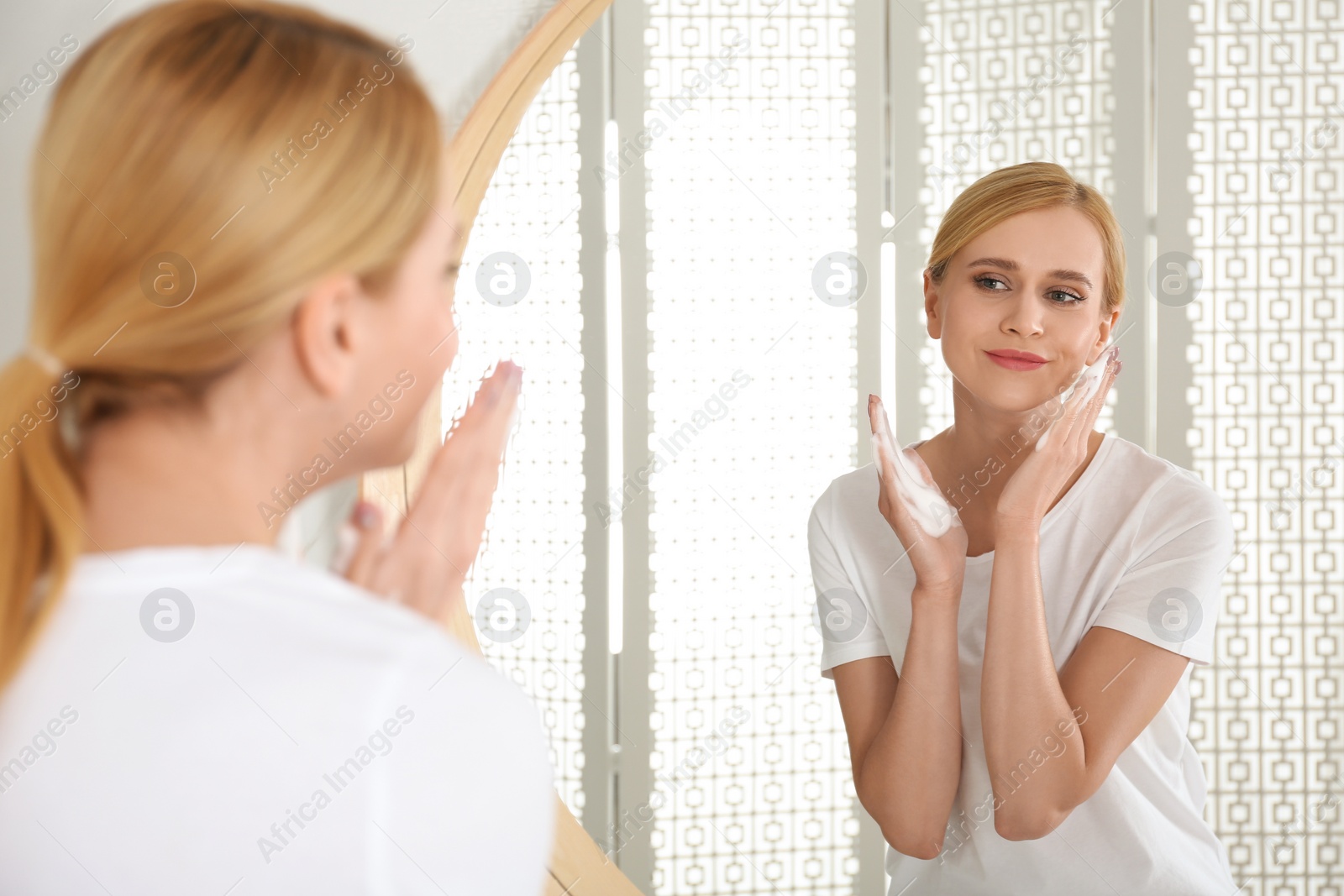 Photo of Happy young woman applying cleansing foam onto face near mirror in bathroom