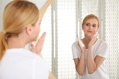 Photo of Happy young woman applying cleansing foam onto face near mirror in bathroom