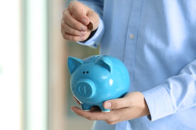 Man putting coin into piggy bank on blurred background, closeup. Money savings