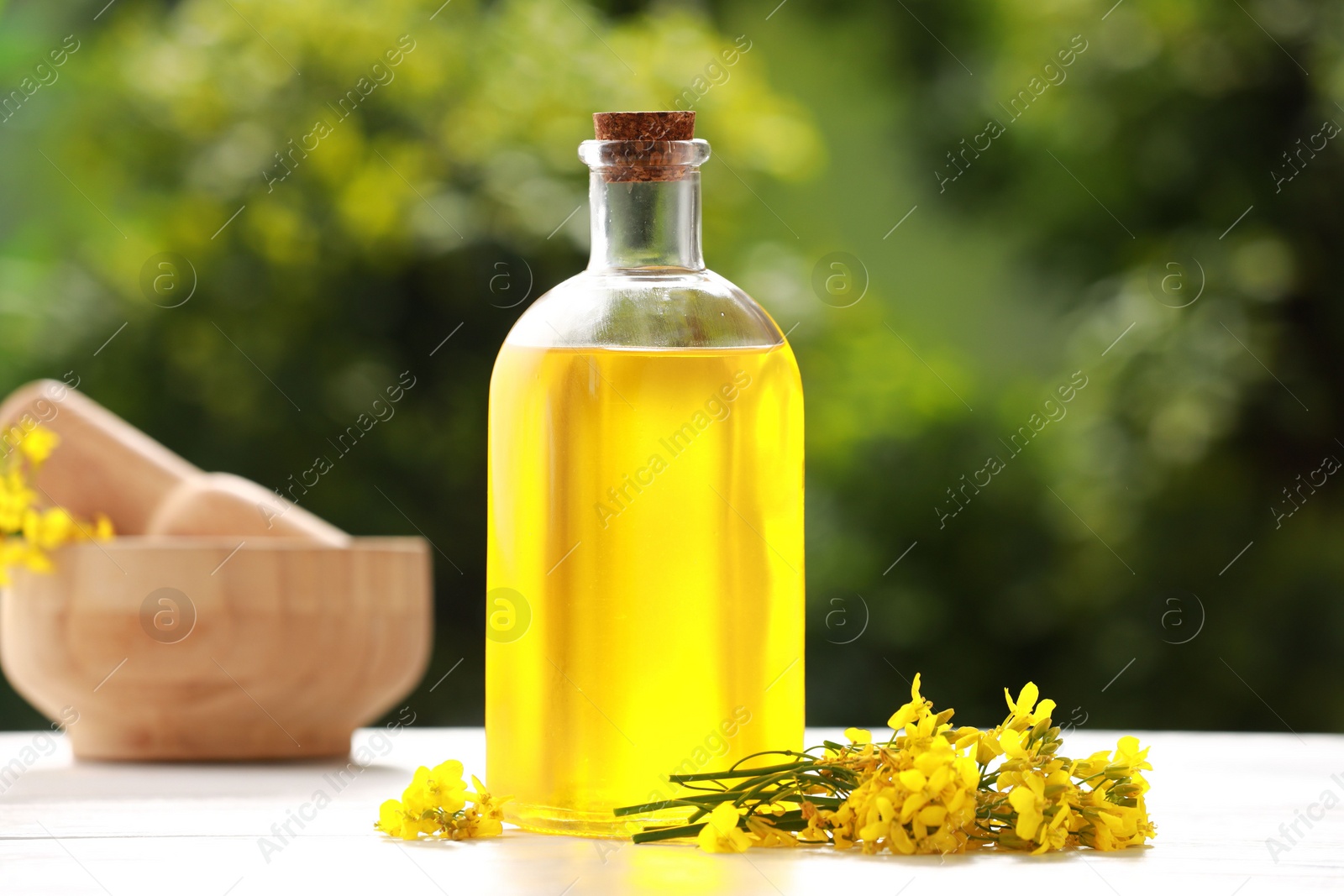 Photo of Rapeseed oil in glass bottle and yellow flowers on white wooden table outdoors