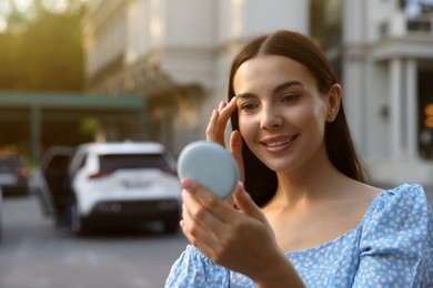 Beautiful young woman looking at herself in cosmetic pocket mirror outdoors. Space for text