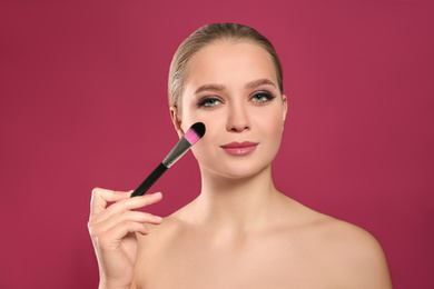 Beautiful woman applying makeup with brush on pink background