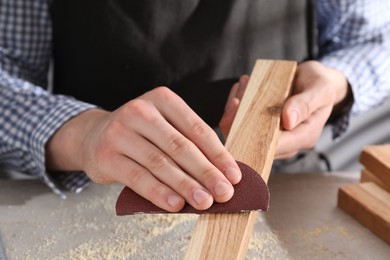 Photo of Man polishing wooden plank with sandpaper at grey table, closeup