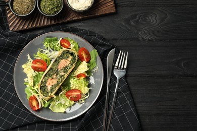 Photo of Piece of tasty strudel with salmon, spinach and salad on black wooden table, flat lay. Space for text