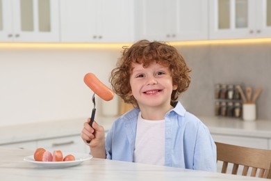 Photo of Cute little boy holding fork with sausages at table in kitchen
