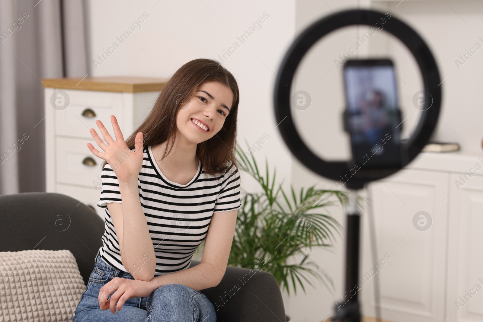 Photo of Smiling teenage blogger waving hello to her subscribers while streaming at home