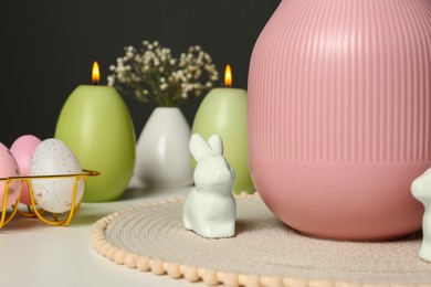 Adorable ceramic bunny figure near pink vase, painted eggs and candles on white table. Easter decor