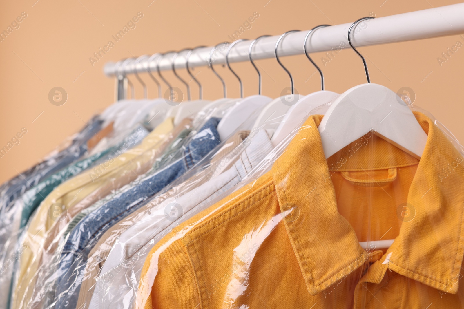 Photo of Dry-cleaning service. Many different clothes in plastic bags hanging on rack against beige background, closeup