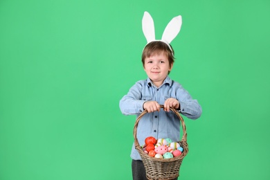 Photo of Cute little boy wearing bunny ears with basket full of dyed Easter eggs on green background, space for text