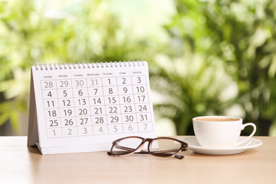 Calendar, glasses and cup of coffee on wooden table against blurred background