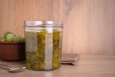 Photo of Jar of delicious gooseberry jam and bowl with fresh berries on wooden table, space for text