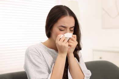 Photo of Sick woman with tissue blowing nose on sofa at home. Cold symptoms