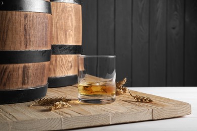Wooden barrels, glass of tasty whiskey and wheat spikes on white table, space for text
