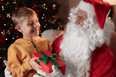 Photo of Little boy taking gift from Santa Claus in room with Christmas tree