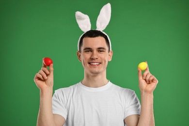 Photo of Easter celebration. Handsome young man with bunny ears holding painted eggs on green background