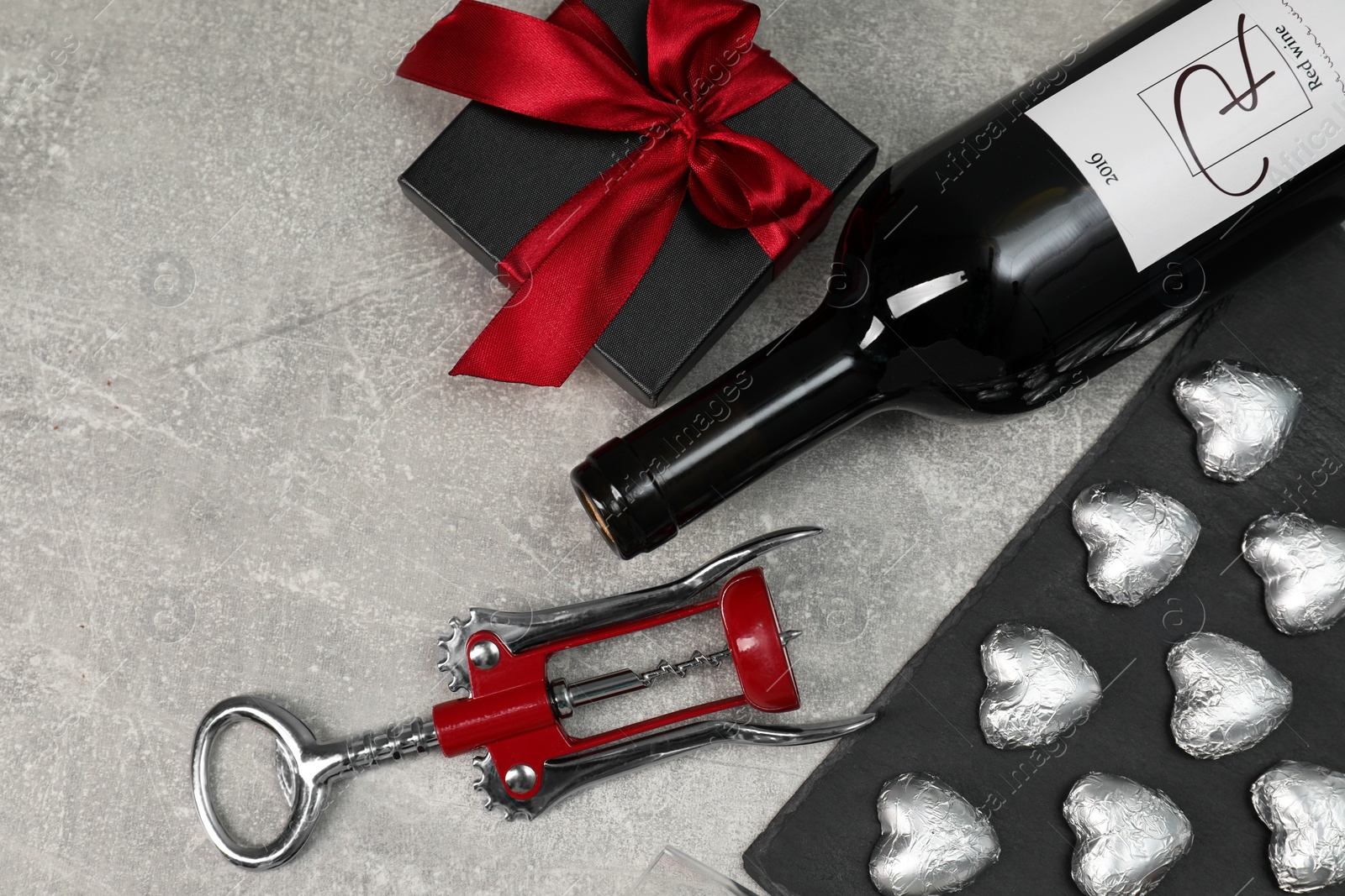 Photo of Bottle of red wine, heart shaped chocolate candies, corkscrew and gift box on light grey textured table, flat lay