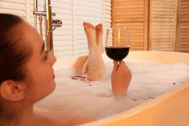 Photo of Woman with glass of wine taking bath in tub with foam and rose petals indoors, closeup