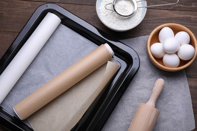 Baking pan with rolls of parchment paper, different ingredients and kitchen tools on wooden table, flat lay