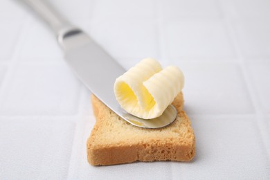 Photo of Tasty butter curl, knife and piece of dry bread on white tiled table, closeup