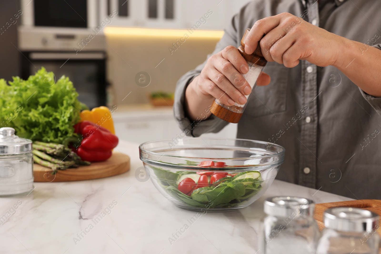 Photo of Cooking process. Man adding salt into bowl of salad at white marble countertop kitchen, closeup